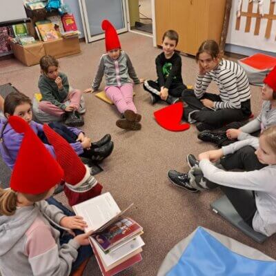 dzieci siedzą w kole na dywanie , dziewczynka w czerwonej czapce czyta książkę. Kliknięcie powoduje powiększenie zdjęcia.