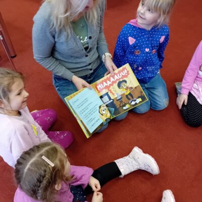 Na dywanie siedzą dzieci wraz z bibliotekarką, która pokazuje im obrazek w książce o kotku.