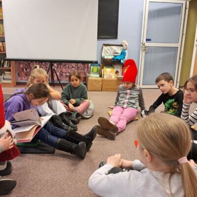 dzieci siedzą w kole na dywanie, jedna dziewczynka trzyma na kolanach czerwonego krasnala, dziewczynka w filetowej bluzie czyta pozostałym dzieciom tekst książki. Kliknięcie powoduje powiększenie zdjęcia.