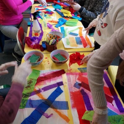 Dzieci siedzące przy stoliku wykonują kompozycje kolorystyczne techniką naklejania pasków bibuły na mokrą kartkę. Kliknięcie powoduje powiększenie zdjęcia.