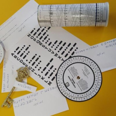 Na stole leżą szyfry i zagadki zapisane alfabetem morsa, piktogramem, kodem Cezara. Kliknięcie powoduje powiększenie zdjęcia.