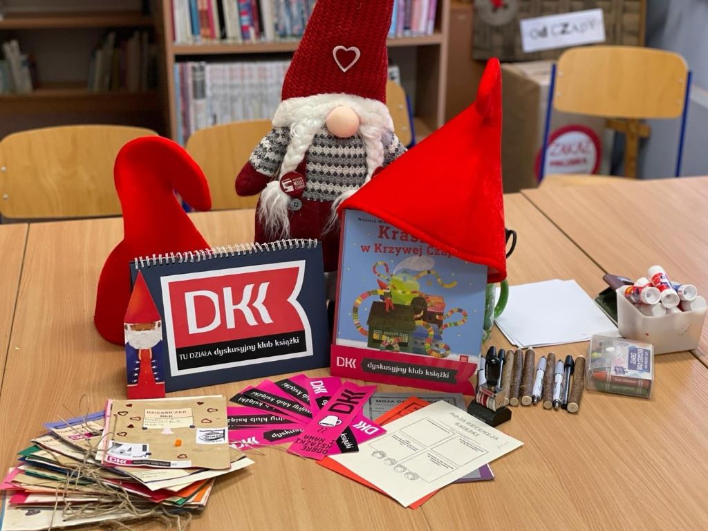 Zdjęcie główne – przedstawia: książkę : „Krasnal w krzywej czapce”, napis DKK, zakładki DKK, dzienniczki DKK ,karty oceny, duży czerwony krasnal, dwie czerwone krasnale czapki, kleje, długopisy i kredki