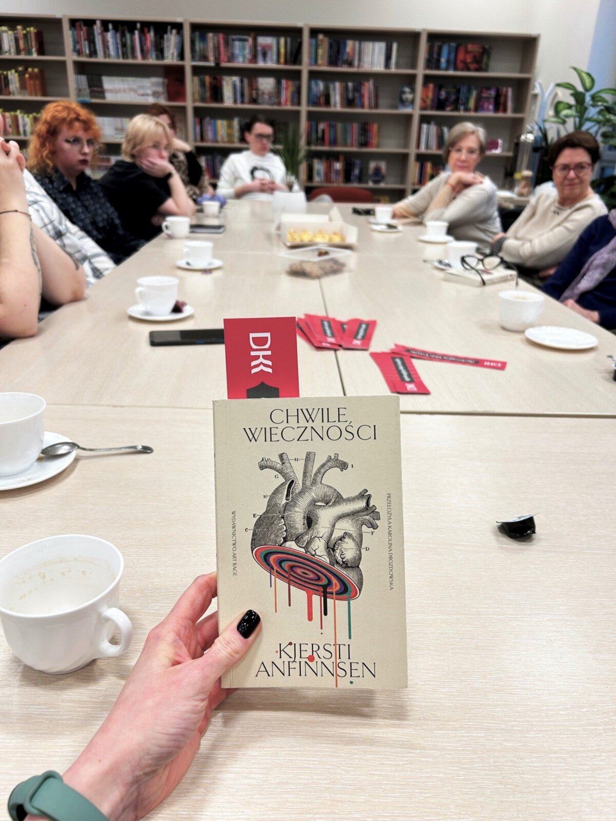 Pokazana jest okładka książki, a w tle widać siedzące przy stole osoby, które biorą udział w spotkaniu DKK.