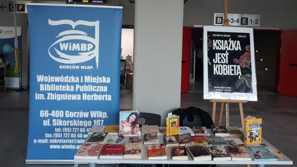 Fotografia przedstawia stół z książkami oraz plakat z hasłem "Książka jest kobietą" i baner z nazwą biblioteki.