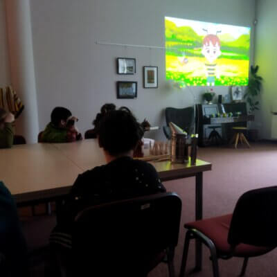 Na ścianie wyświetlana jest prezentacja multimedialna, kilkoro dzieci siedzących przy stole przy zgaszonym świetle. Kliknięcie w obrazek spowoduje powiększenie.