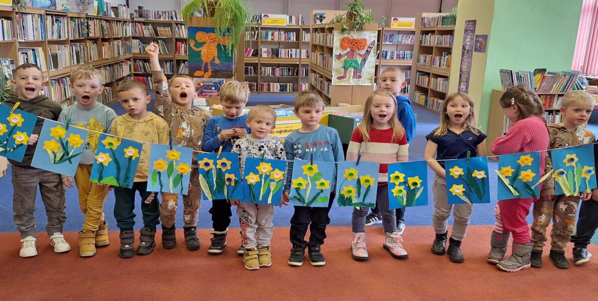Grupa dzieci przedszkolnych z radosnym okrzykiem „Wiosna!” prezentuje swoje prace plastyczne. Kartki z żonkilami rozwieszone na sznurku.