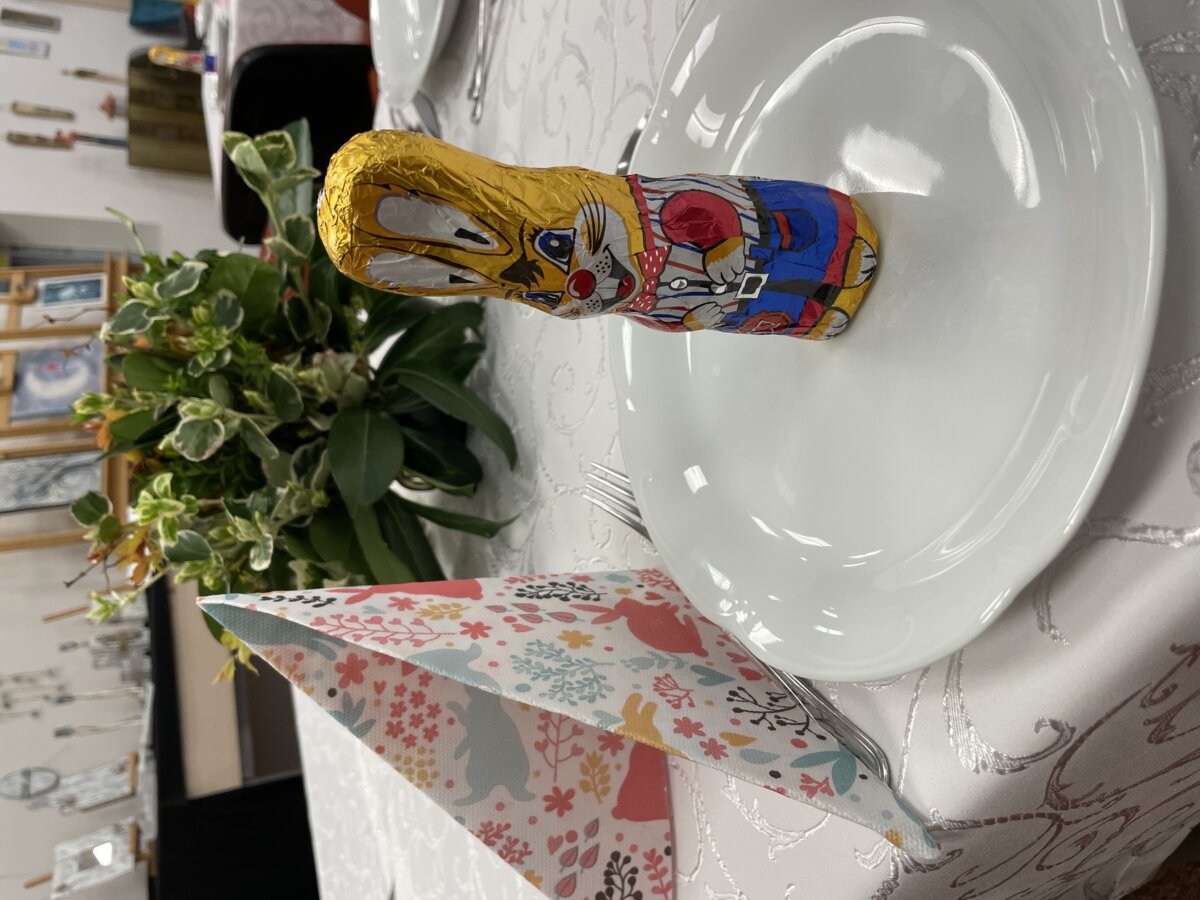 Fragment stołu, na białym obrusie biały talerzyk, na którym stoi czekoladowy zajączek w ozdobnym złotku, obok zielona dekoracja w formie bukietu.