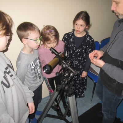 Prezentacja teleskopu słonecznego; czworo dzieci stoi naprzeciw prowadzącego warsztaty mężczyzny: troje patrzy na teleskop, jedno coś mówi. Kliknięcie w obrazek spowoduje powiększenie.