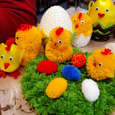 Ręcznie robione prace jednej z uczestniczek warsztatów. Kurczaki siedzące na trawie obok kolorowych jajek. Wszystko wykonane z włóczki. W tle białe ozdobne jajo z perełkami oraz kurczaki z innych materiałów. Kliknięcie powoduje powiększenie zdjęcia.