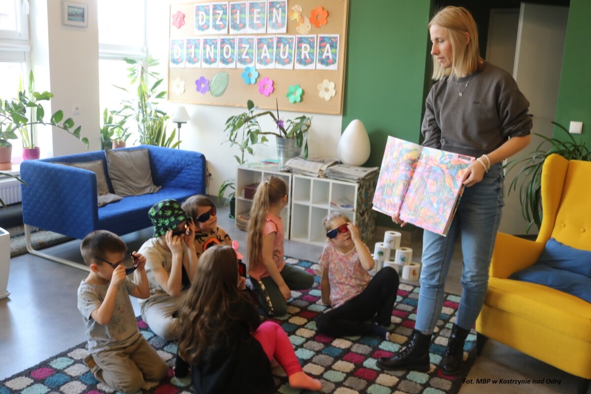 Kobieta prezentuje książkę dzieciom siedzącym w okularach 3D na dywanie.