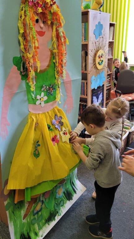 Dzieci ozdabiają zrobionymi przez siebie papierowymi, kolorowymi kwiatami postać Pani Wiosny, wykonaną za pomocą farb i bibuły na dużej, przypiętej do regału kartce