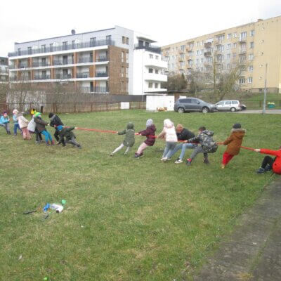 Na rozległym trawniku, niedaleko chodnika dzieci bawią się w przeciąganie grubej czerwonej liny; pojedynek jest zaciekły, część zawodników aż przysiada z wysiłku na trawie; w tle widać niskie czteropiętrowe bloki mieszkalne. Kliknięcie w obrazek spowoduje powiększenie.