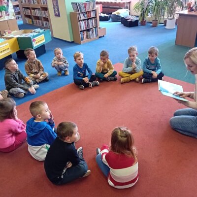 Dzieci siedzą w kółku na dywanie i słuchają wiersza czytanego przez bibliotekarkę. Kliknięcie w obrazek spowoduje powiększenie.