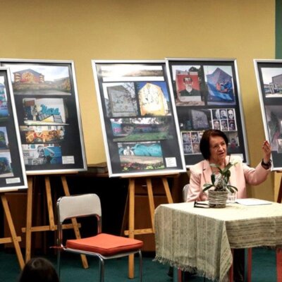 Prowadząca spotkanie Maria Karbowska siedzi przy stoliku. Opowiada o swoich spacerach. Za nią stoją sztalugi z wystawą. Kliknięcie w obrazek spowoduje powiększenie.