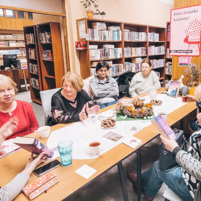 Dyskusyjny Klub Książki. Siedem kobiet siedzi przy stole w bibliotece. Toczy się dyskusja. Niektóre kobiety gestykulują i się uśmiechają. Kliknięcie powoduje powiększenie zdjęcia.