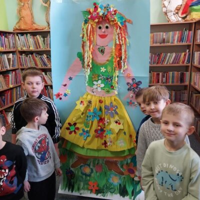 Grupa chłopców stojąca na tle postaci Pani Wiosny, namalowanej na dużej kartce, w sukience wykonanej z bibuły, ozdobionej kolorowymi kwiatami. Klikniecie powoduje powiększenie zdjęcia.