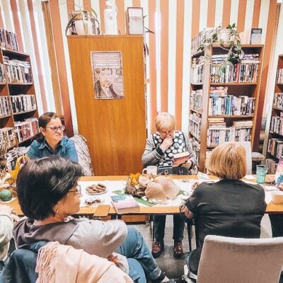 Wnętrze biblioteki. Trwa spotkanie Dyskusyjnego Klubu Książki. Siedem kobiet rozmawia. W tle regały z książkami. Na pierwszym planie długi stół. Kliknięcie powoduje powiększenie zdjęcia.