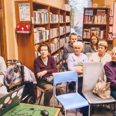 Wnętrze biblioteki. Na widowni siedzi siedem osób: sześć kobiet i jeden mężczyzna. Przy stoliku po lewej siedzi kobieta prowadząca spotkanie. Uczestnicy słuchają opowieści. Kliknięcie powoduje powiększenie zdjęcia.