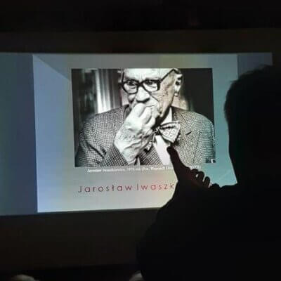 W zaciemnionym pomieszczeniu ekran z wyświetlonym slajdem przedstawiającym zdjęcie Jarosław Iwaszkiewicza. Na tle ekranu widoczny fragment ciemnej sylwetki prowadzącego. Kliknięcie powoduje powiększenie zdjęcia.