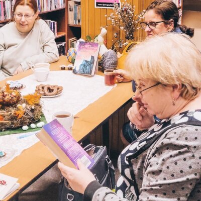 Biblioteka. Filia nr 8. Zbliżenie na trzy kobiety słuchające rozmowy o książce. Jedna z nich trzyma lekturę w dłoni i czyta opis na tylnej okładce. Na stole ciastka i wielkanocne ozdoby. Kliknięcie powoduje powiększenie zdjęcia.