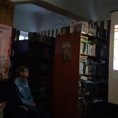 Zaciemnione wnętrze biblioteki. Po prawej widoczny rzutnik z wyświetlonym slajdem. Po lewej część osób przybyłych na wydarzenie. Uczestnicy patrzą na ekran. Kliknięcie powoduje powiększenie zdjęcia.
