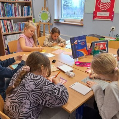 Dzieci siedzą przy stole i wypełniają dzienniczki DKK. Kliknięcie powoduje powiększenie zdjęcia.
