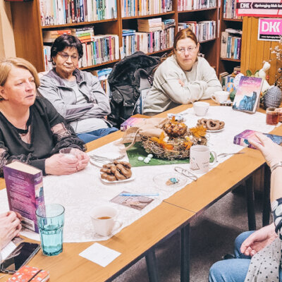 Wnętrze biblioteki. Trwa Dyskusyjny Klub Książki. Siedem kobiet siedzących przy stole rozmawia o omawianej książce. W tle regały z książkami. Kobieta po prawej gestykuluje. Inne na nią patrzą. Kliknięcie powoduje powiększenie zdjęcia.