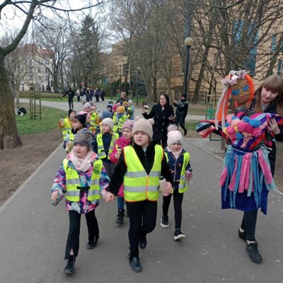 Dzieci wraz z opiekunami oraz Paniami Bibliotekarkami spacerują po parku, wesoło śpiewając i niosąc marzannę. Kliknięcie powoduje powiększenie zdjęcia.