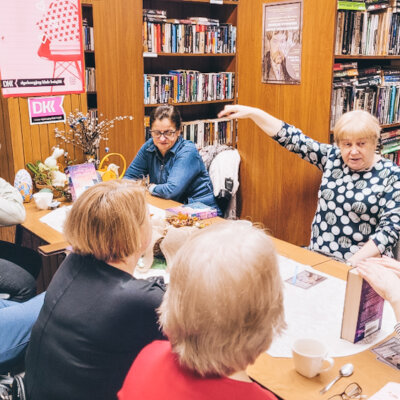 Biblioteka. Siedem kobiet uczestniczy w spotkaniu Dyskusyjnego Klubu Książki. Jedna z kobiet wskazuje na coś ręką i toczy swoją opowieść. Inne kobiety przysłuchują się historii. Kliknięcie powoduje powiększenie zdjęcia.