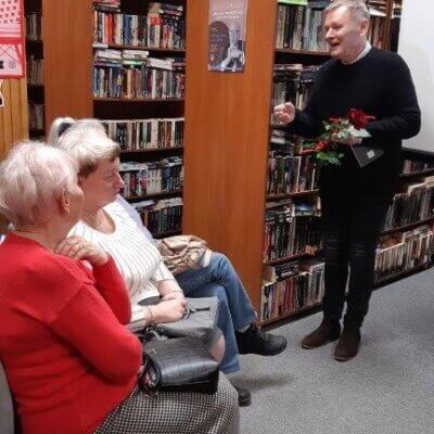 Prowadzący wieczór literacki stojąc zwraca się do osób zgromadzonych w bibliotece. W ręku trzyma różę i tomik wierszy. Kliknięcie powoduje powiększenie zdjęcia.