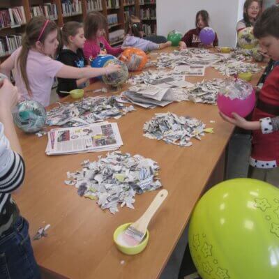 Na dużym stole w czytelni biblioteki rozłożone są w dużych ilościach skrawki papieru gazetowego, kolorowe balony i miseczki z szerokimi pędzlami; dookoła stołu stoi dziesięcioro dzieci i oklejają balony masą papierową. Kliknięcie w obrazek spowoduje powiększenie.