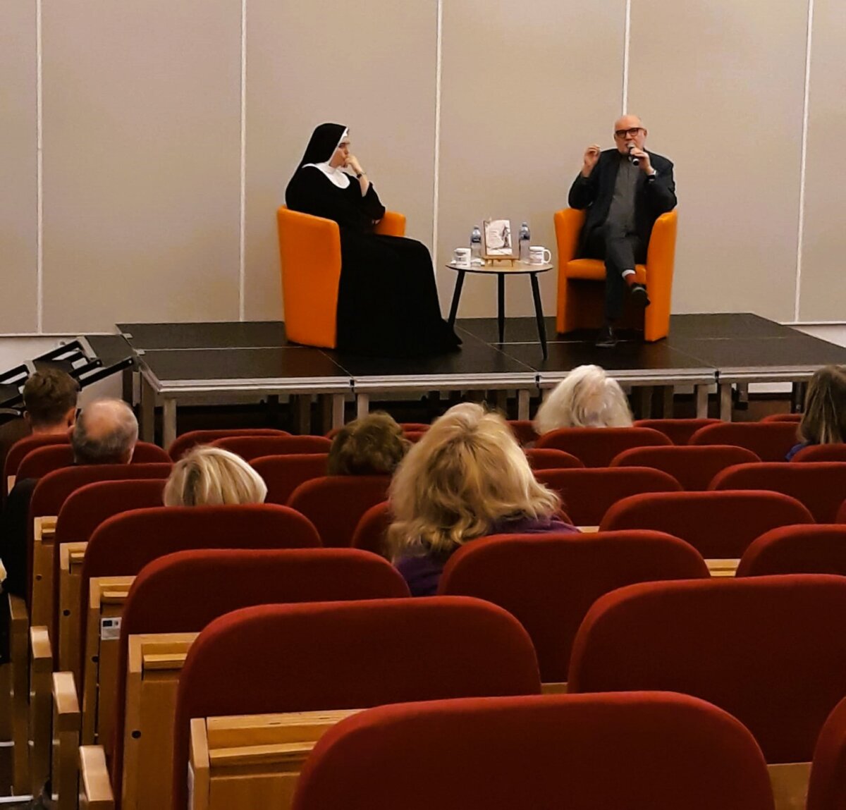 fotografia prezentuje siedzących na fotelach: księdza i siostrę zakonną, rozmawiających o promowanej książce. Na pierwszym planie widać siedzących uczestników spotkania.