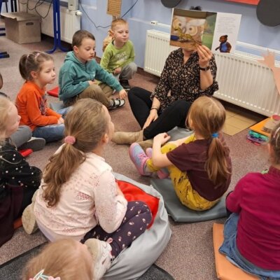 Dzieci siedzą na dywanie, bibliotekarka pokazuje dzieciom ilustracje. Kliknięcie powoduje powiększenie zdjęcia.