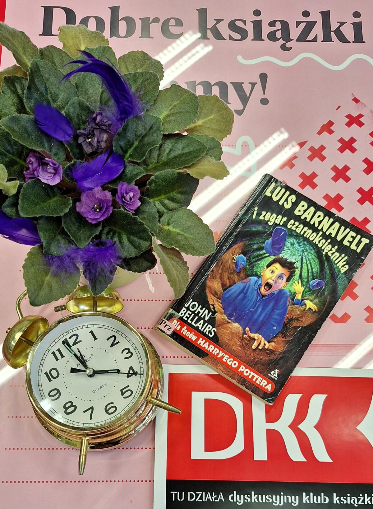 Na plakacie DKK stoi fiołek w doniczce, pod nim leży złoty zegar, a po prawej stronie książka „Luis Barnavelt i zegar czarnoksiężnika”.