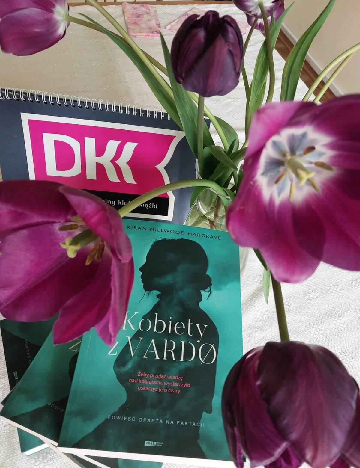 Na kremowej serwecie wazon z fioletowymi tulipanami wśród nich w zielono-czarnej kolorystyce książka Kiran Millwood Hargrave, pt.: „Kobiety z Vardo”, fioletowy emblemat Dyskusyjnego Klubu Książki.