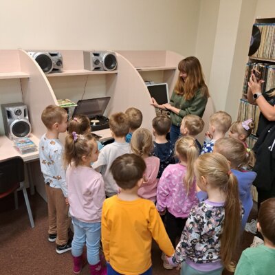 Zdjęcie przedstawia grupę kolorowo ubranych przedszkolaków i bibliotekarkę, która prezentuje dzieciom sposób działania gramofonu. W tle widać biurka ze sprzętem elektronicznym i fragment regału z płytami gramofonowymi. Kliknięcie powoduje powiększenie zdjęcia.