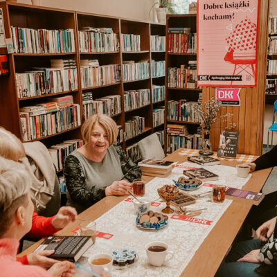 Trwa Dyskusyjny Klub Książki. Kobiety siedzą przy stole w bibliotece. Toczy się dyskusja. Jedna z kobiet się uśmiecha. Inna czyta opis książki z tylnej okładki. Na stole leży omawiana książka. Kliknięcie powoduje powiększenie zdjęcia.