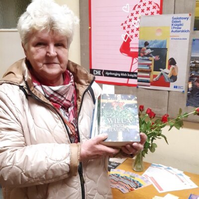 Lokalna autorka Halina Elżbieta Daszkiewicz pozuje z książką na tle plakatu i czerwonych róż. Kliknięcie w obrazek spowoduje powiększenie.