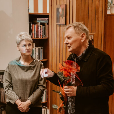 Wnętrze biblioteki. Kierowiczka Filii nr 8 wraz z prowadzącym spotkanie literackie. Janusz Dreczka gestykuluje prawą ręką. W lewej trzyma czerwoną różę. Kliknięcie w obrazek spowoduje powiększenie.