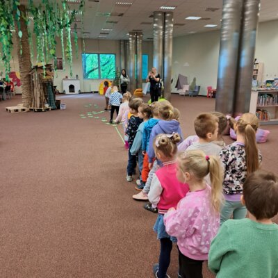 Zdjęcie przedstawia kolorowo ubranych przedszkolaków, którzy stoją w rzędzie. W oddali dwóch przedszkolaków skacze po zielonych śladach zostawionych przez żabki. W tle widać także przedszkolanki obserwujące dzieci oraz drzewo wykonane z materiałów plastycznych. Kliknięcie powoduje powiększenie zdjęcia.
