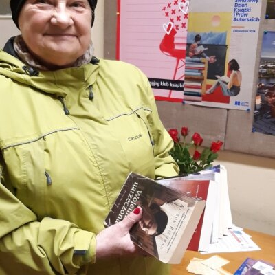 Uśmiechnięta kobieta w jasnej kurtce stoi na tle plakatu i czerwonych róż. W rękach trzyma wypożyczone książki. Kliknięcie w obrazek spowoduje powiększenie.