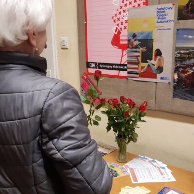Kobieta w szarej kurtce stoi tyłem. W ręce trzyma czerwoną różę. W tle plakat promujący akcję oraz czerwone róże. Kliknięcie w obrazek spowoduje powiększenie.