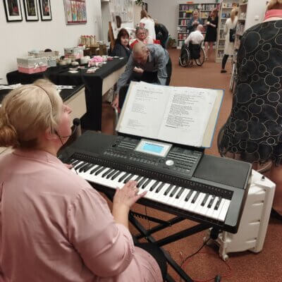 Goście zwiedzaja wystawę, na pierwszym planie kobieta gra na instrumencie klawiszowym. Kliknięcie powoduje powiększenie zdjęcia.
