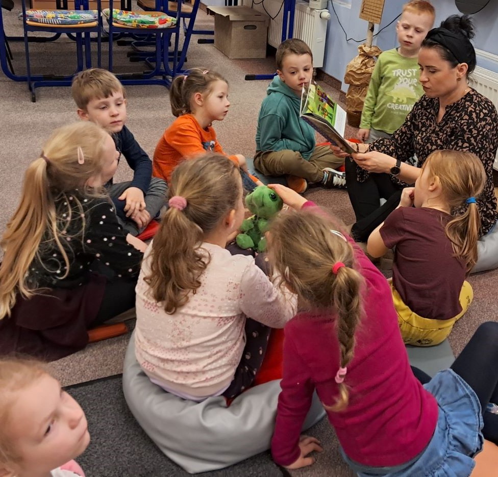 dzieci siedzą na dywanie, bibliotekarka czyta dzieciom książkę, dwie dziewczynki trzymają na kolanach maskotką – żółwia