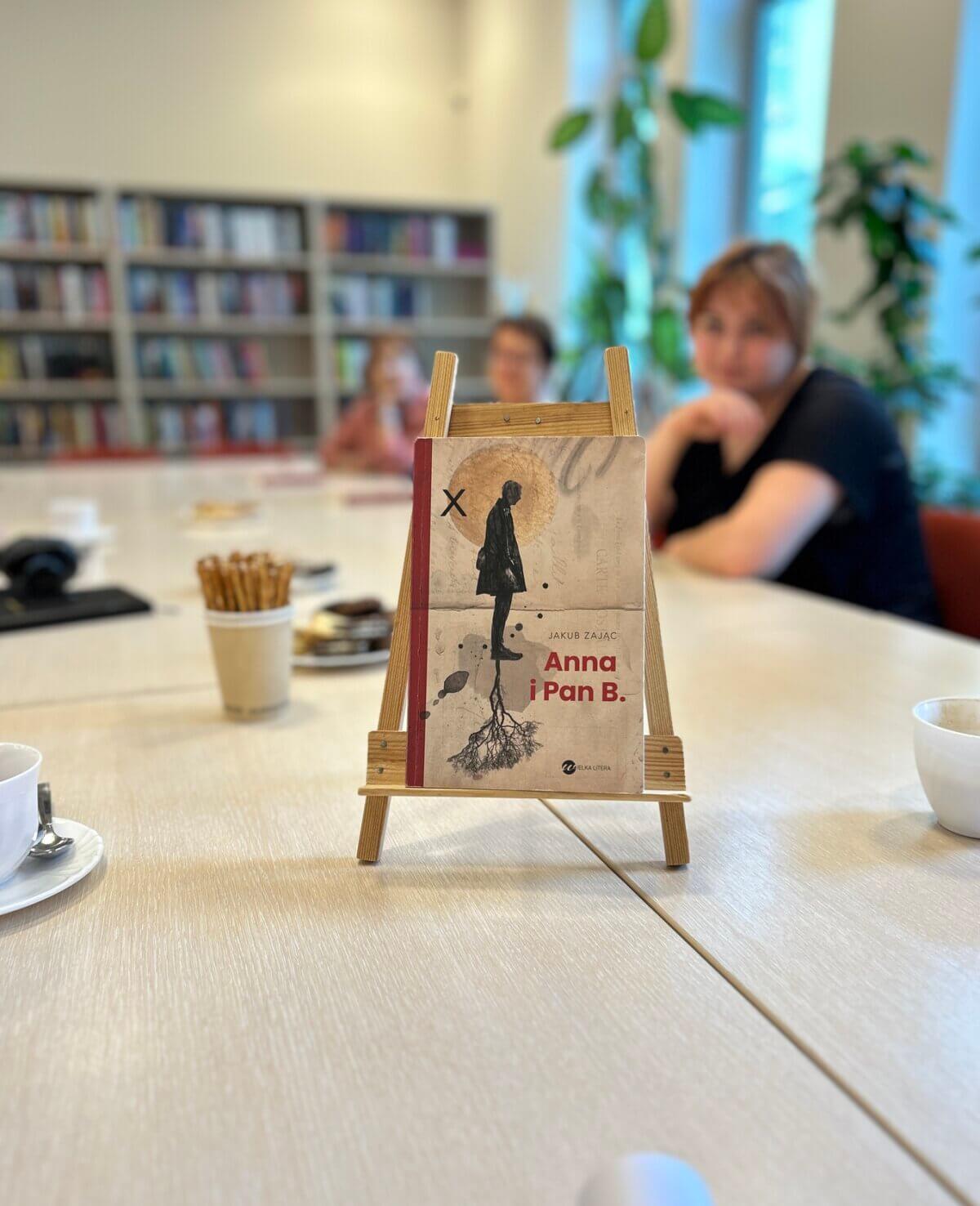 Na zdjęciu widać książkę „Anna i Pan B.” której autorem jest Jakub Zając. Książka jest oparta o stojak i stoi na stole. W tle widać rozmyte uczestniczki.